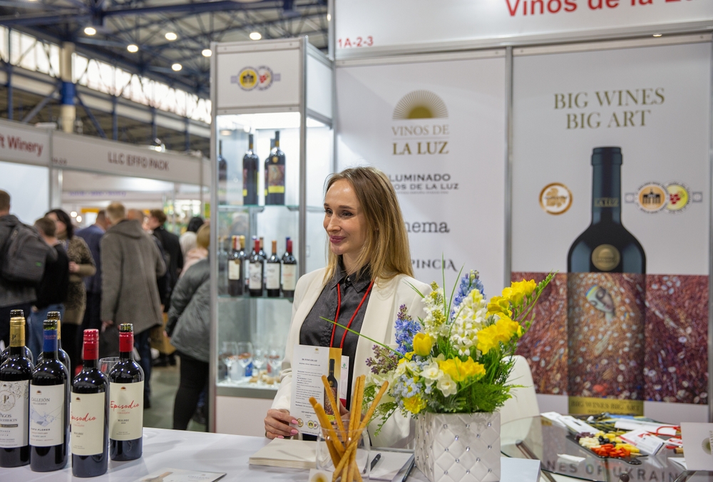 A wine trade booth at a trade show, with a hostess and an array of wine bottles on display.
